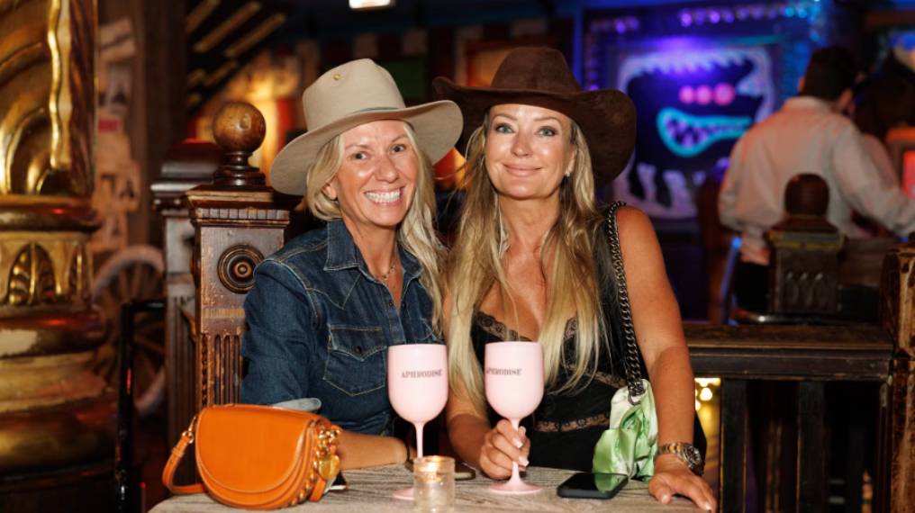 Two women wearing cowboy hats smiling holding a glass of Aphrodise