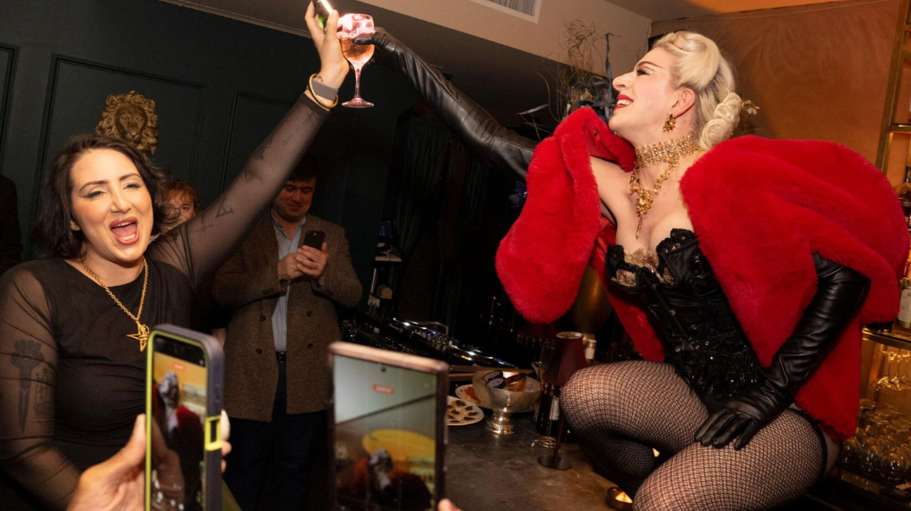 Person kneeling on top of a table, cheersing glasses with another person