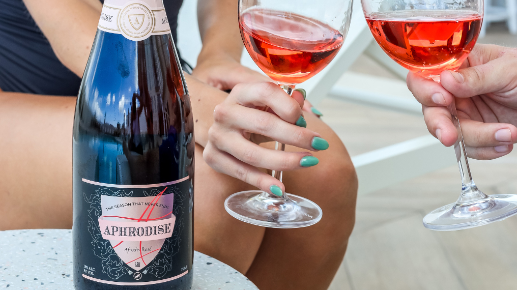 A bottle of Aphrodise at the forefront with two people cheersing glasses of Aphrodise wine in the background