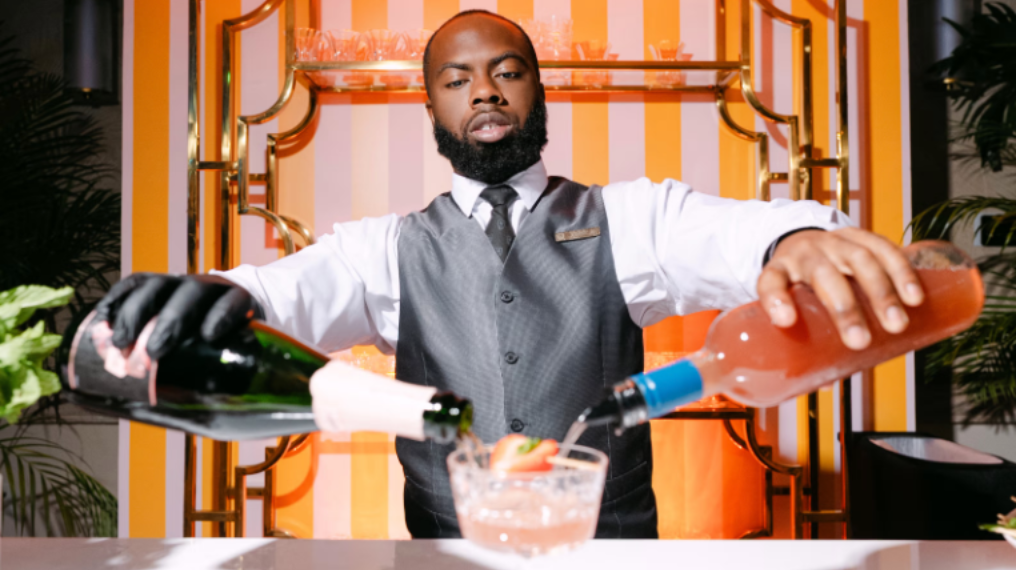 Waiter holding bottles in each hand, pouring to mix a cocktail in a glass in front